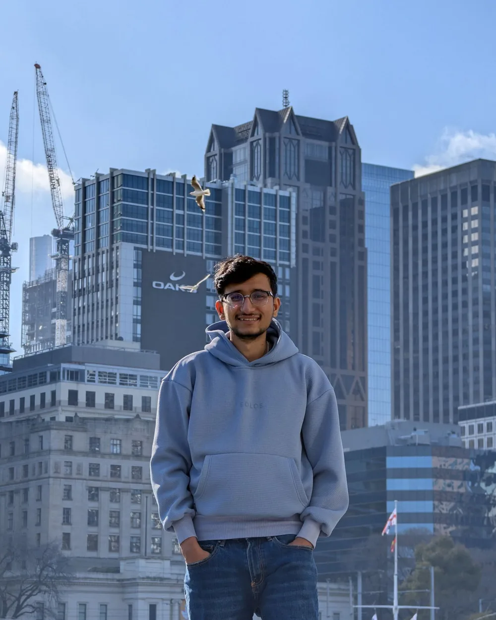 Arun Sapkota in a grey hoodie, smiling, with Melbourne buildings and construction cranes behind him